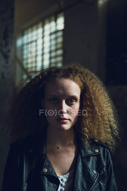 Frontansicht einer jungen kaukasischen Frau mit lockigem Haar, die eine Lederjacke trägt, während sie in einem dunklen und leeren Lagerhaus aufmerksam in die Kamera blickt — Stockfoto