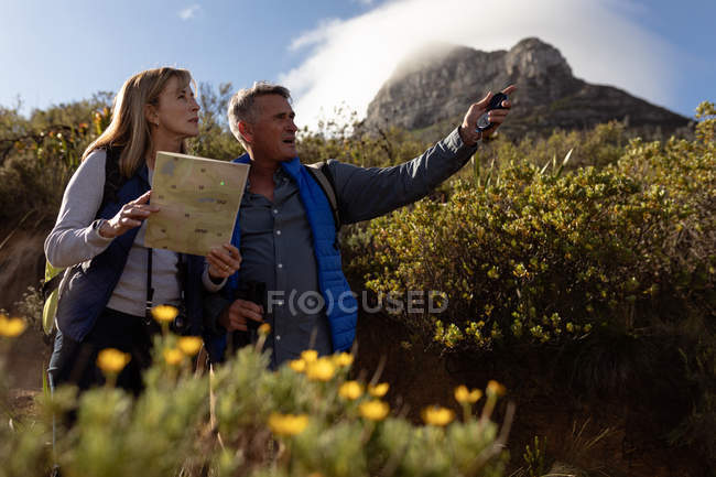 Vue latérale rapprochée d'une femme et d'un homme caucasiens matures, la femme tenant une carte et l'homme pointant vers la distance lors d'une randonnée à la campagne, un sommet de montagne et un nuage en arrière-plan — Photo de stock