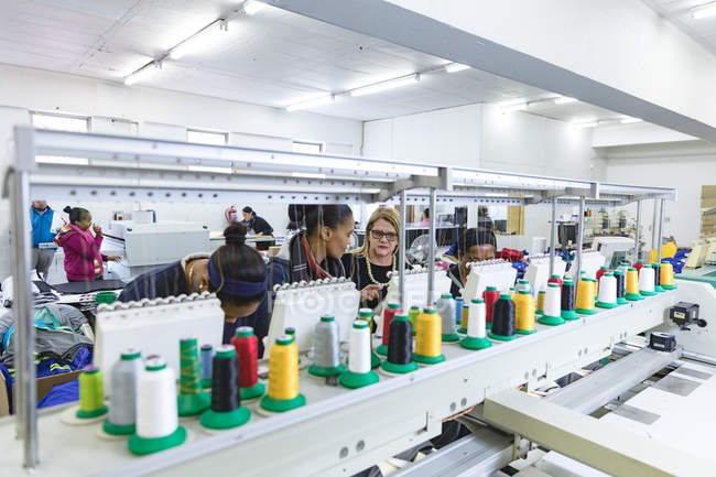 Vista frontal de un grupo diverso de colegas mujeres que trabajan detrás de una fila de máquinas de coser automatizadas en una fábrica de ropa deportiva . - foto de stock