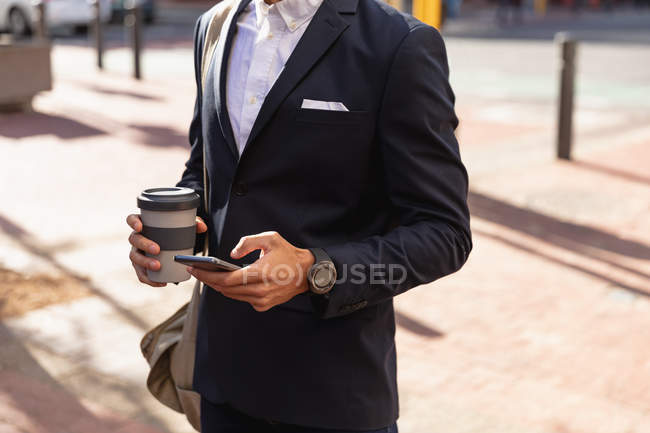 Vista frontal sección media del hombre con una chaqueta usando su teléfono inteligente y sosteniendo un café para llevar, de pie en una calle de la ciudad. Nómada digital en movimiento . - foto de stock