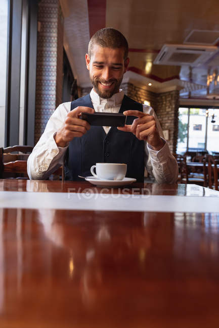 Vista frontal de cerca de un joven caucásico sonriente usando su teléfono inteligente sentado en una mesa con una taza de café dentro de un café. Nómada digital en movimiento . - foto de stock