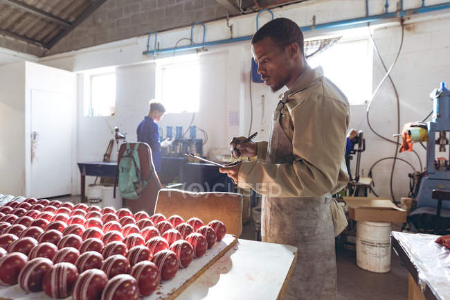 Vista lateral en medio de un joven afroamericano sosteniendo un portapapeles y escribiendo mientras revisa filas de bolas de cricket al final de la línea de producción en una fábrica de equipos deportivos . - foto de stock