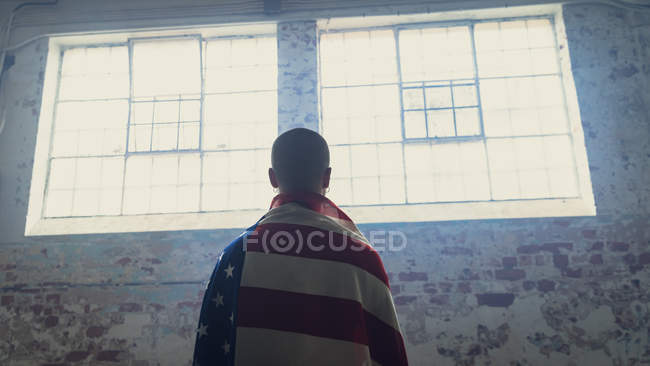 Visão traseira de um jovem com costas viradas contra a câmera e uma bandeira americana sobre ombros dentro de um armazém vazio — Fotografia de Stock