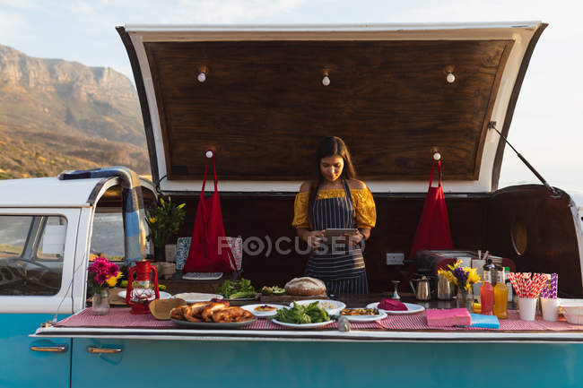 Vue de face d'une jeune femme métisse utilisant une tablette dans un van à toit ouvert offrant une gamme de plats à emporter à vendre — Photo de stock
