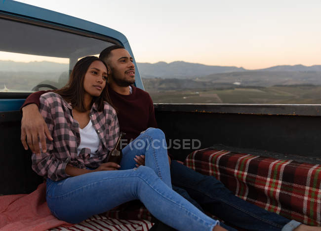 Vista lateral de una joven pareja mixta sentada en la parte trasera de su camioneta, abrazando y disfrutando de la vista al atardecer durante una parada en un viaje por carretera . - foto de stock