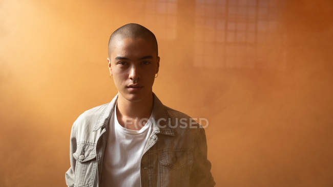Front view of a young Hispanic-American man wearing a grey leather jacket over a white shirt looking intently at the camera inside an empty warehouse with orange smoke — Stock Photo