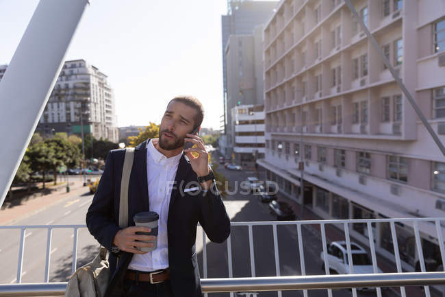 Вид спереди на молодого кавказца, держащего кофе на вынос и разговаривающего по смартфону, стоящего на дорожке по городской дороге. Цифровая реклама на ходу . — стоковое фото