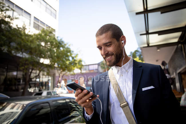 Vista frontal de cerca de un joven caucásico sonriente usando un teléfono inteligente y usando auriculares en una calle de la ciudad. Nómada digital en movimiento . - foto de stock