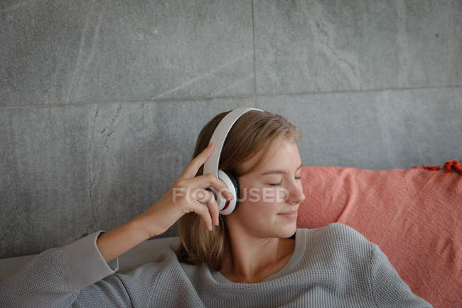 Nahaufnahme einer jungen kaukasischen Frau, die mit weißen Kopfhörern auf einem Sofa sitzt und mit geschlossenen Augen Musik hört. — Stockfoto