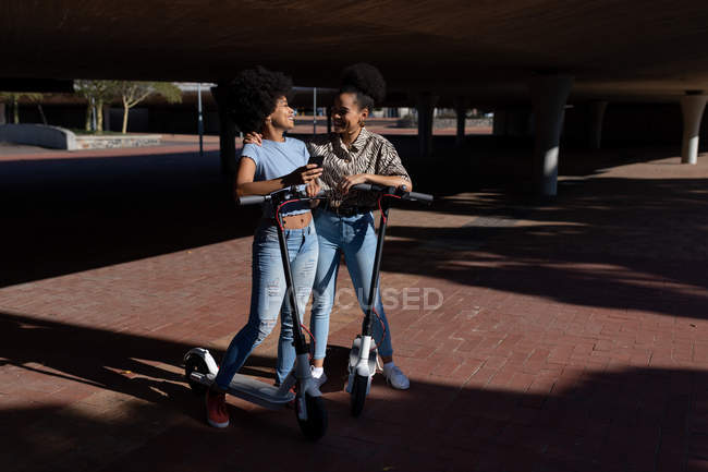 Вид спереди на двух молодых взрослых смешанных расовых сестер, стоящих с электрическими скутерами в городском парке с помощью смартфона. Одна обнимает другую, и они смотрят друг на друга, улыбаясь — стоковое фото