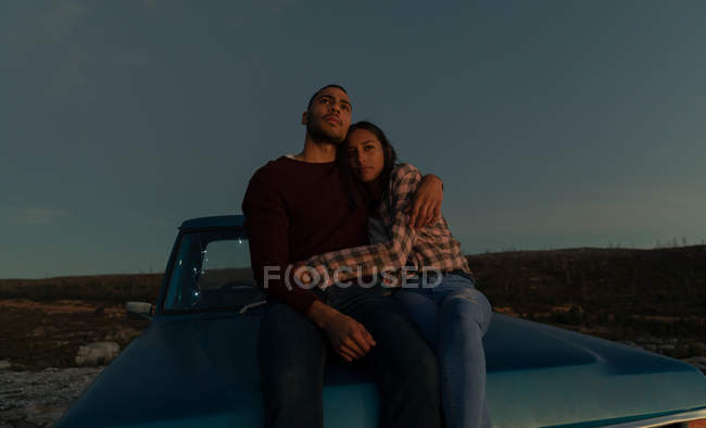 Vista frontal de um jovem casal misto sentado na parte de trás de seu caminhão pick-up, abraçando e apreciando a vista ao entardecer, durante uma parada em uma viagem de carro . — Fotografia de Stock