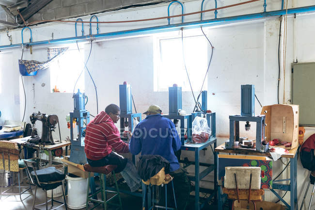 Visão traseira de dois jovens mestiços trabalhadores do sexo masculino sentados, falando e operando máquinas em uma fábrica que faz bolas de críquete . — Fotografia de Stock