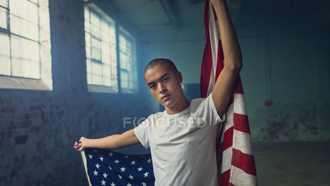 Front view of a young Hispanic-American man wearing a plain white shirt holding an American flag looking intently at the camera inside an empty warehouse — Stock Photo