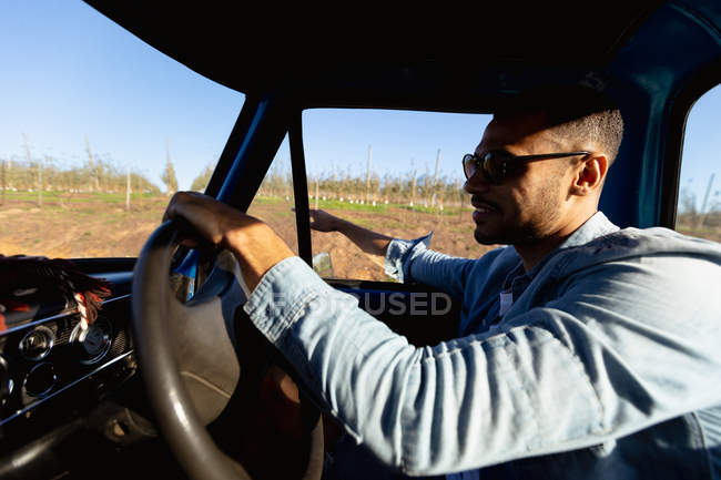 Vista lateral de cerca de un joven y sonriente hombre de raza mixta sentado al volante de una camioneta durante un viaje por carretera - foto de stock