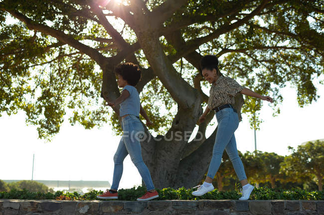 Vue de côté gros plan de deux jeunes sœurs souriantes adultes métis marchant le long d'un mur dans un parc urbain, avec leurs scooters électriques garés en dessous d'eux — Photo de stock