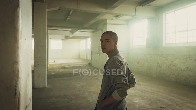 Side view of a young Hispanic-American man with piercings wearing a grey jacket with hands in pockets looking intently at the camera inside an empty warehouse — Stock Photo