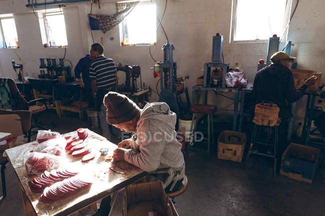 Elevated side view of a young African American man wearing a hat sitting at a workbench working with cut out red leather shapes in a workshop at a factory making cricket balls, in the background colleagues are working — Stock Photo