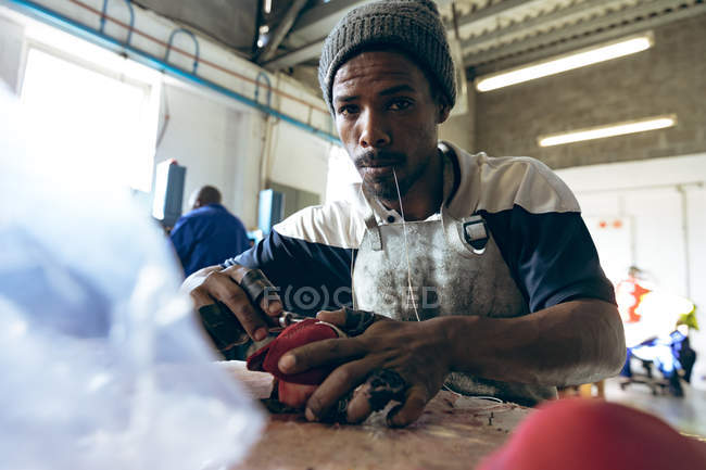 Vue de face gros plan d'un jeune Afro-Américain assis sur un établi avec du fil dans la bouche, cousant des formes de cuir rouge dans une usine fabriquant des balles de cricket et regardant vers la caméra, les matériaux et l'équipement au premier plan . — Photo de stock
