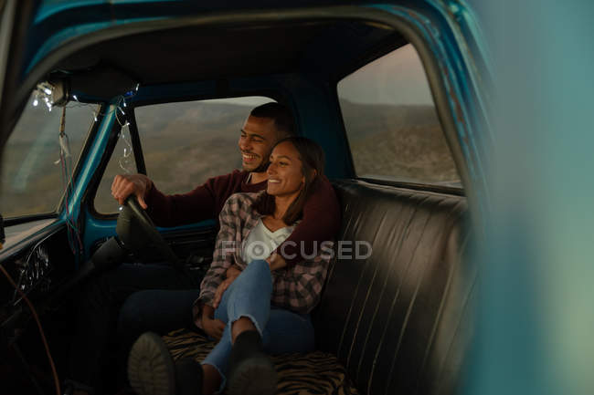 Vista lateral de una joven pareja mixta sentada en su camioneta sonriendo y abrazándose al atardecer, durante una parada en un viaje por carretera. Están sentados en los asientos delanteros y el interior del coche está iluminado con luces de cuerda . - foto de stock