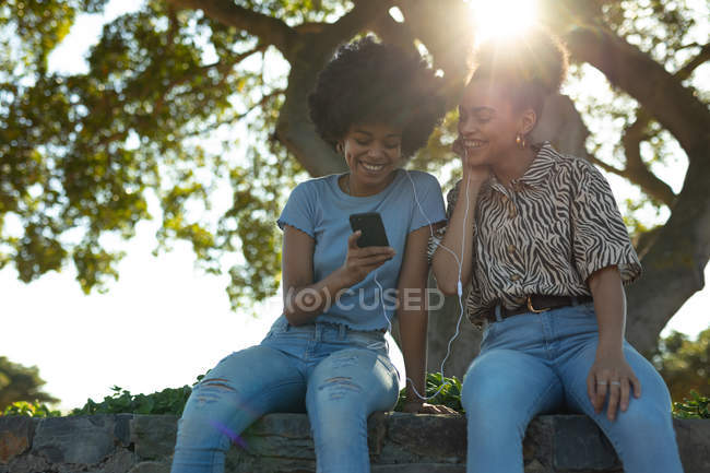 Vista frontale da vicino di due giovani sorridenti sorelle di razza mista adulte sedute su un muro in un parco urbano, che usano uno smartphone e condividono gli auricolari per ascoltare musica, retroilluminate con il bagliore delle lenti — Foto stock