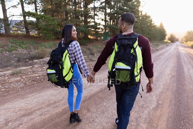 Vista trasera de una joven pareja de raza mixta con mochilas, tomados de la mano y mirándose mientras caminan por un camino rural, el camino se extiende por delante de ellos con un entorno rural en el fondo - foto de stock