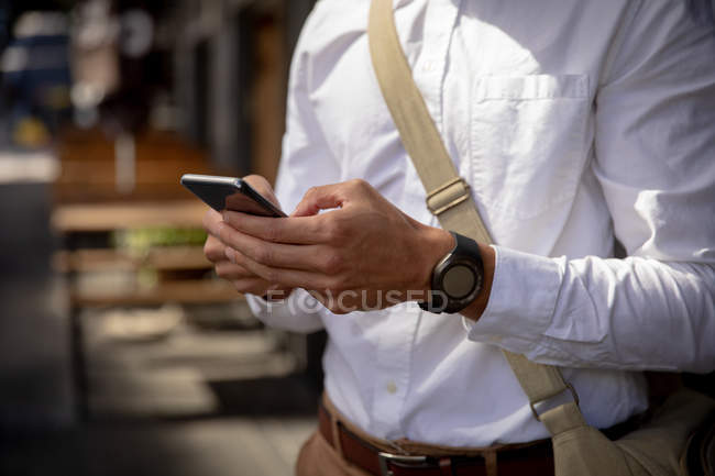 Seitenansicht eines Mannes mit Umhängetasche und Smartphone, der auf dem Bürgersteig einer Stadtstraße steht. Digitaler Nomade unterwegs. — Stockfoto