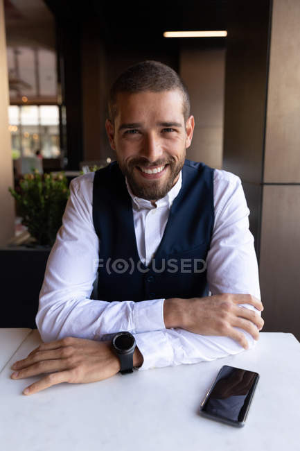 Ritratto ravvicinato di un giovane caucasico sorridente seduto a un tavolo all'interno di un caffè, con lo smartphone sul tavolo accanto a lui. Nomade digitale in movimento . — Foto stock