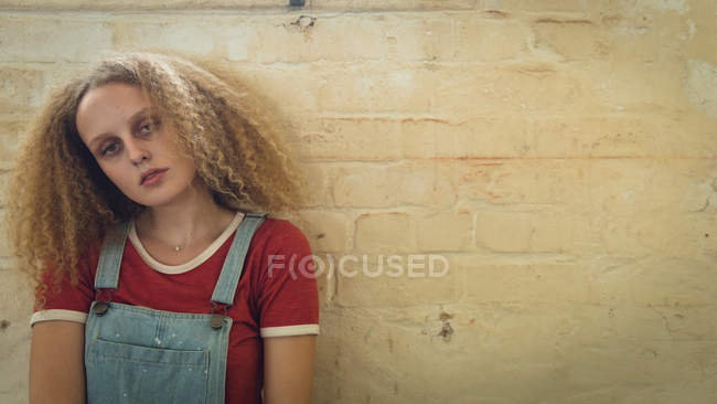 Vista frontal de una joven mujer caucásica con el pelo rizado mirando atentamente a la cámara con una pared de ladrillo en el fondo - foto de stock