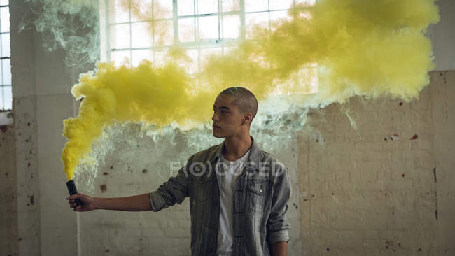 Frontansicht eines jungen hispanisch-amerikanischen Mannes, der eine graue Jacke über einem weißen Hemd trägt und von der Kamera wegschaut, während er eine Rauchmaschine in der Hand hält, die gelben Rauch in einer leeren Lagerhalle produziert — Stockfoto