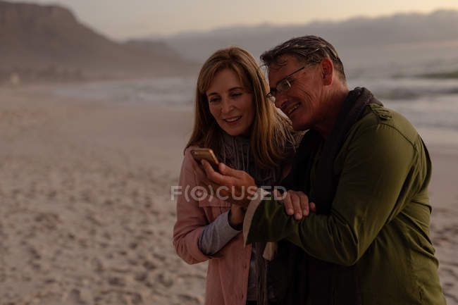Вид сбоку на зрелого кавказца: мужчина и женщина улыбаются и смотрят на смартфон на пляже у моря на закате — стоковое фото