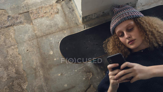 High angle view of a young Caucasian woman with curly hair wearing a black shirt and beanie lying on the floor with head on a skateboard while using a mobile phone inside an empty warehouse — Stock Photo