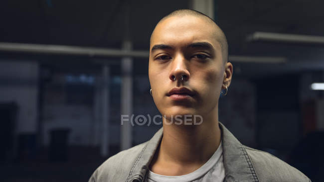 Close up view of a young Hispanic-American man with piercings wearing a grey jacket over a white shirt while standing inside a dark and empty warehouse and looking away from the camera — Stock Photo