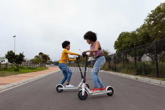 Vista lateral de dos hermanas adultas jóvenes de raza mixta montadas en scooters eléctricos en un camino tranquilo - foto de stock