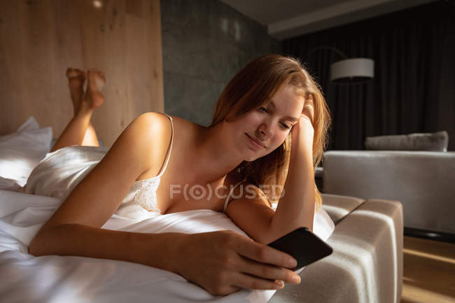 Close up de jovem mulher loira caucasiana deitada na cama usando smartphone. Ela está relaxando e praticando o autocuidado . — Fotografia de Stock