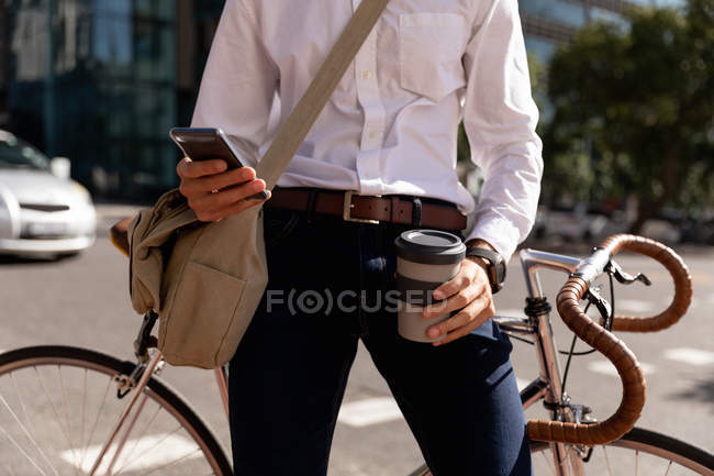 Вид спереди: мужчина, держащий в руках кофе и пользующийся смартфоном, опирается на велосипед на городской улице. Цифровая реклама на ходу . — стоковое фото