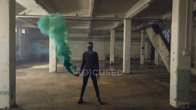 Vorderansicht eines Mannes mit halb verdecktem Gesicht, der lange Ärmel trägt und eine Rauchmaschine in der Hand hält, die grünen Rauch in einer leeren Lagerhalle produziert — Stockfoto