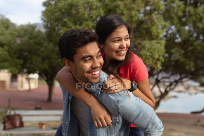 Front view close up of a smiling young mixed race couple having fun piggybacking outside in the sun and looking away, with trees and the sea in the background — Stock Photo