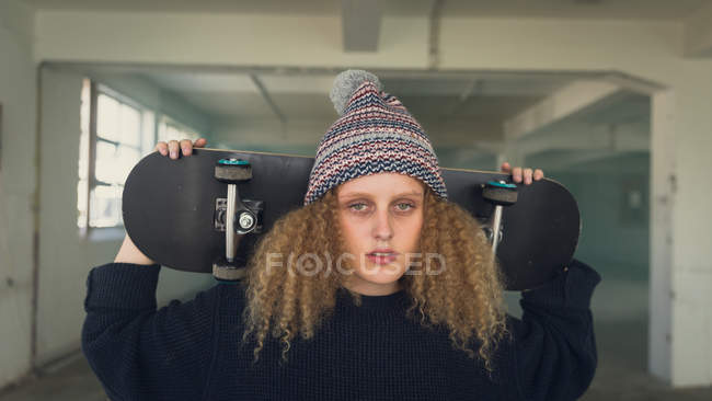 Frontansicht einer jungen kaukasischen Frau mit lockigem Haar, die schwarze lange Ärmel und Mütze trägt, während sie ein Skateboard über der Schulter hält und in eine leere Lagerhalle aufmerksam in die Kamera blickt — Stockfoto