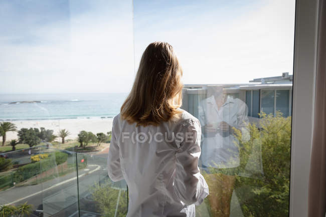Vista posteriore primo piano di una giovane donna caucasica che indossa una camicia bianca in piedi vicino a una finestra che tiene una tazza di caffè e guarda fuori, mare e spiaggia sullo sfondo . — Foto stock