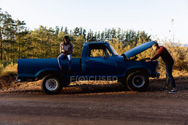 Side view of a young mixed couple having a breakdown with their pick-up truck during a road trip. The woman is sitting on the back of the truck using a smartphone, while the man has the hood open and is trying to fix the truck — Stock Photo