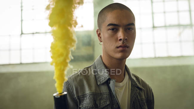 Vista frontal de un joven hispano-americano con una chaqueta gris sobre una camisa blanca mirando atentamente a la cámara mientras sostiene una máquina de humo que produce humo amarillo dentro de un almacén vacío - foto de stock