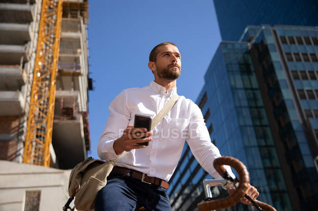 Vista frontal de perto de um jovem caucasiano usando um smartphone, sentado em sua bicicleta e olhando para longe em uma rua da cidade. Digital Nomad em movimento . — Fotografia de Stock