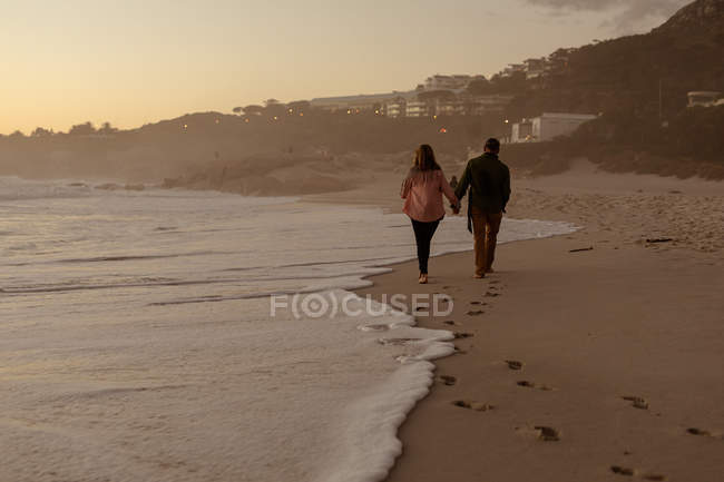 Вид збоку зрілого Кавказького чоловіка і жінки тримає руки і ходить уздовж пляжу біля моря на заході сонця — стокове фото