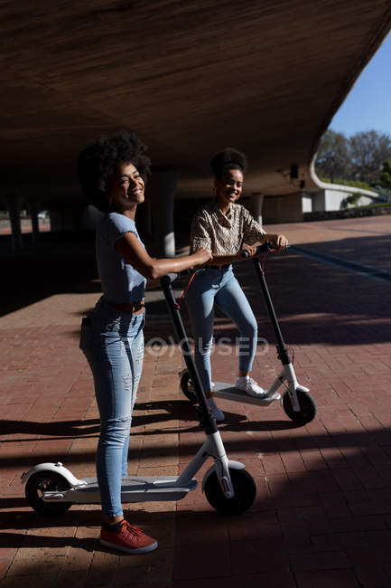 Vista lateral de dos hermanas adultas jóvenes de raza mixta de pie con scooters eléctricos en un parque urbano, mirando a la cámara y sonriendo - foto de stock