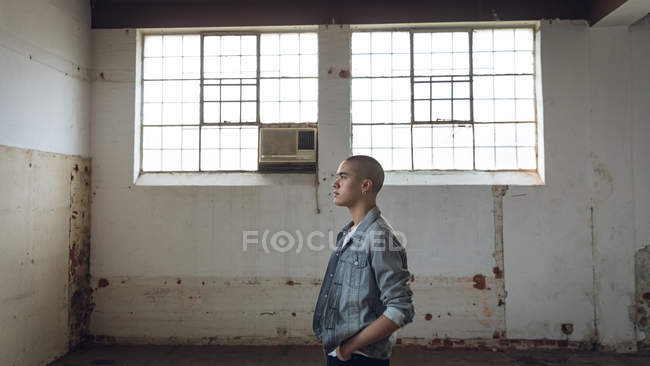 Seitenansicht eines jungen spanisch-amerikanischen Mannes mit Piercings, der eine graue Jacke und eine Hand in der Tasche in einer leeren Lagerhalle trägt — Stockfoto
