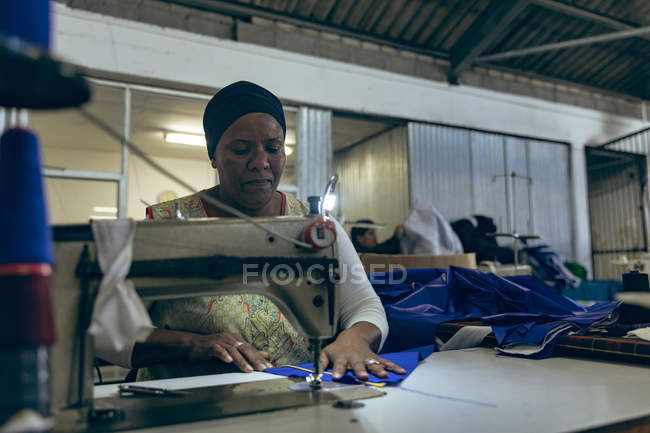 Вид спереду крупним планом жінка середнього віку змішаної раси, яка сидить і керує швейною машиною на заводі спортивного одягу . — стокове фото