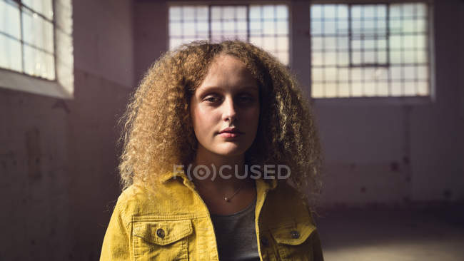 Frontansicht einer jungen kaukasischen Frau mit lockigem Haar, die eine gelbe Jacke über einem grauen Hemd trägt und in einer leeren Lagerhalle aufmerksam in die Kamera blickt. — Stockfoto