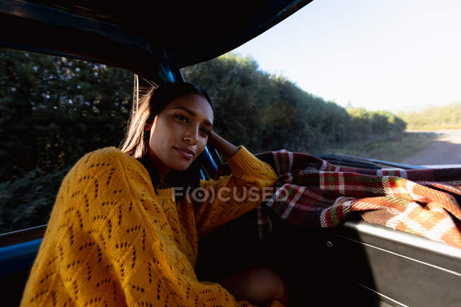 Primer plano de una joven mujer de raza mixta sentada en el asiento delantero del pasajero de una camioneta apoyada en el salpicadero y sonriendo durante un viaje por carretera - foto de stock