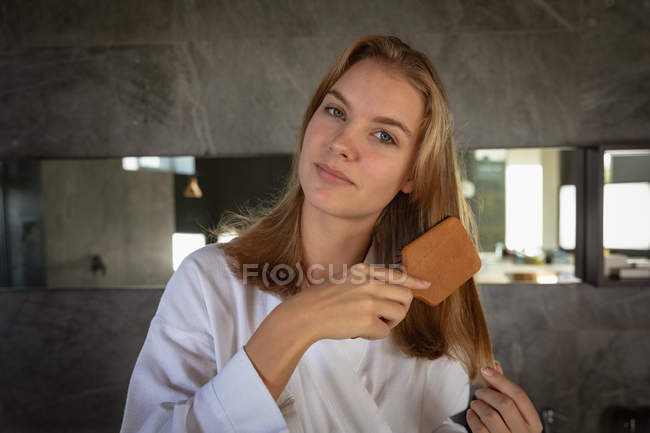 Ritratto da vicino di una giovane donna caucasica che indossa un accappatoio spazzolandosi i capelli, guardando dritto alla macchina fotografica in un bagno moderno . — Foto stock