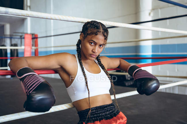 Female boxer with boxing gloves leaning on ropes and looking at camera in boxing ring. Strong female fighter in boxing gym training hard. — Stock Photo